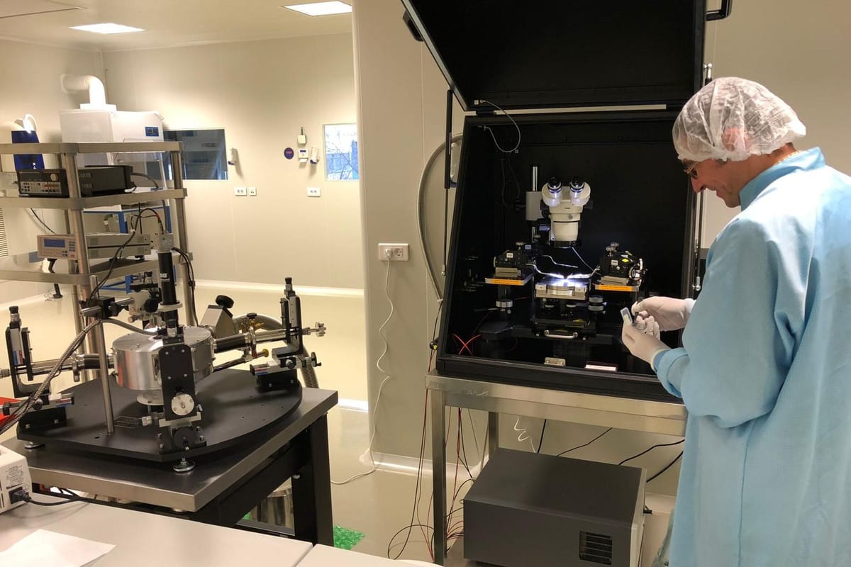 A researcher in a cleanroom gown holding a sample beside a probe station inside a light-tight enclosure.