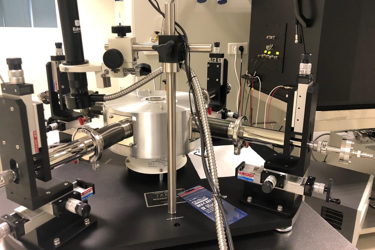 A cryogenic probe station on an optical table, with micropositioners and a stereo microscope arranged around a central cryostat.